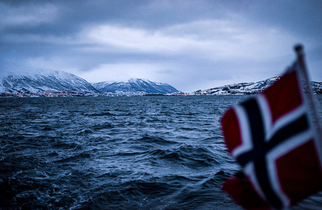 A photo with a Norwegian flag in the foreground, overlooking a fjord and mountains in the background.