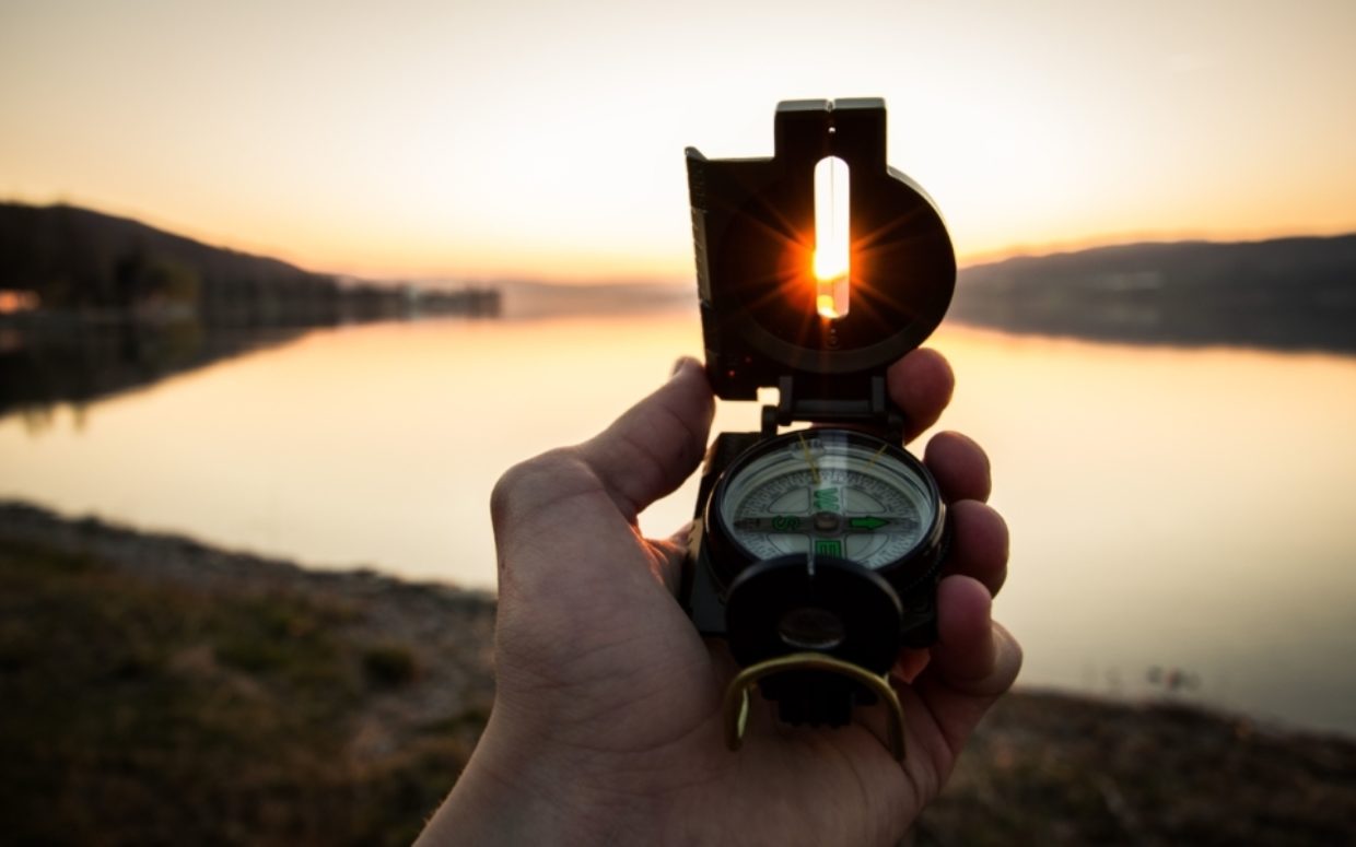 A hand holding a compass overlooking a lake in the sunset